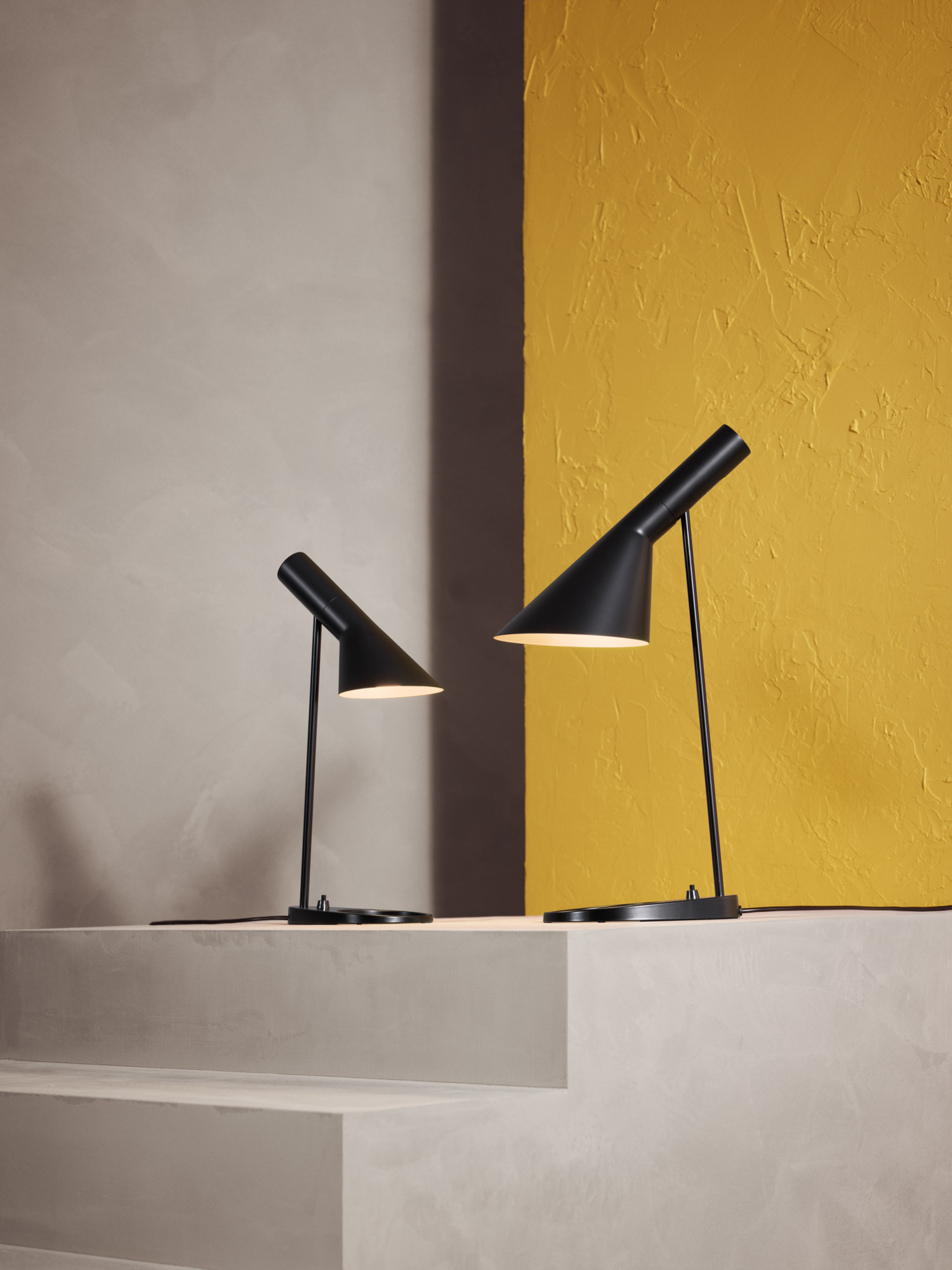 AJ Mini Table lamp & AJ Table lamp in black on concrete steps with a yellow wall backdrop, © 2025 Louis Poulsen.